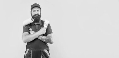 serious bearded man renovator in work clothes hold paint roller and brush on yellow background.