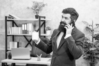 Happy professional man in suit talking on cellphone while drinking tea in office, phone.