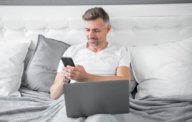 smiling mature man chatting on phone and working on laptop in bed.