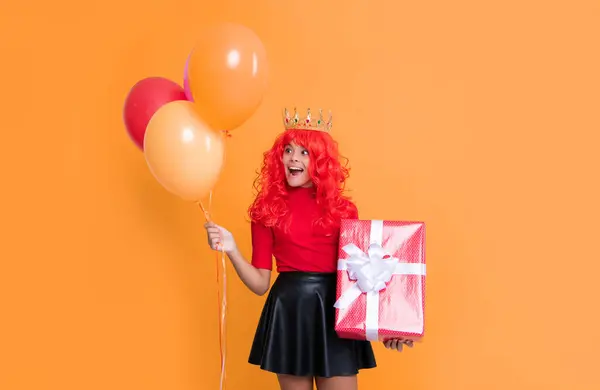 Stock image shocked child in crown with present box and party balloon on yellow background.