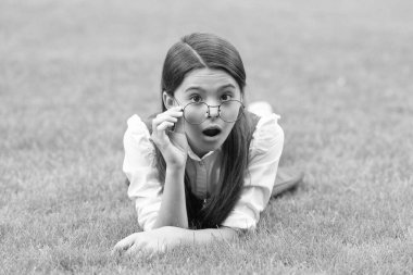Portrait of surprised teen girl adjusting glasses lying on grass after school, surprise. Teen girl with surprised face.