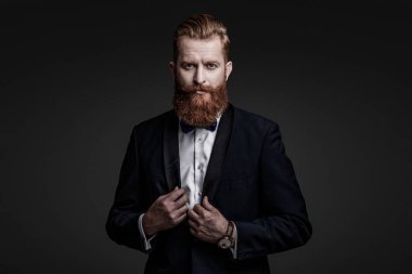 Şık smokin papyonu. Sakallı damat kendinden emin ve hırslı hissediyor. Smokinli erkek giysili adam Griler 'de izole edilmiş. Smokinli adam resmi erkek giyim bölümünde. Yakışıklı sakallı adam smokin giyer. Damat..
