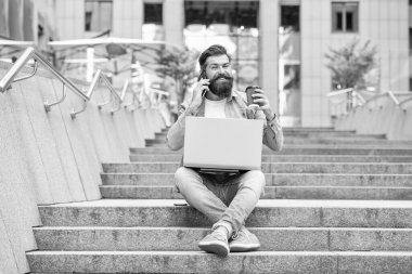 Cep telefonuyla konuşan, merdivenlerde kahve içen mutlu bir adam. Gülümseyen adam dışarıdaki bilgisayarı kullanarak cep telefonu görüşmesi yapıyor. Mobil iletişim. Telefon görüşmesi. Cep telefonu çağrısı.