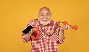 cheerful senior man with retro telephone and modern smartphone on yellow background.