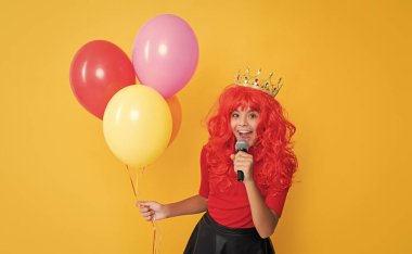 glad child in crown with microphone and party balloon on yellow background.