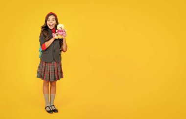 Sarı arka planda sırt çantası ve oyuncağı olan okul üniformalı hayret verici çocuk, kopya alanı, Eylül.