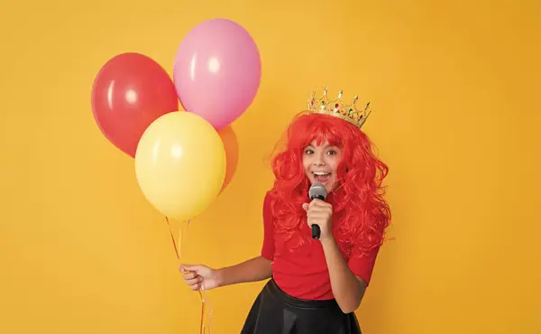 stock image glad child in crown with microphone and party balloon on yellow background.