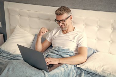 Gözlüklü mutlu bir adam laptopla yatakta çalışıyor..