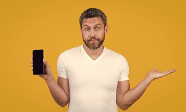 Kafası karışmış bir adam telefonda uygulama gösteriyor. Adam cep telefonunda kopyalama alanı olan bir uygulama gösteriyor. Sarı arka planda izole edilmiş telefon uygulaması gösteren bir adam. Stüdyoda telefon uygulaması gösteren adam.