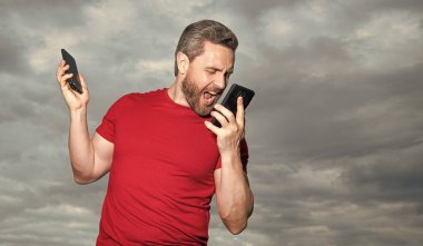 Bağıran kızgın adamın fotoğrafı telefonda konuşuluyor. Adamın gökyüzü geçmişinde telefon konuşmaları var. Adam dışarıda telefonla konuşuyor. Adam telefonda konuşuyor. Kırmızı tişört giyiyor..
