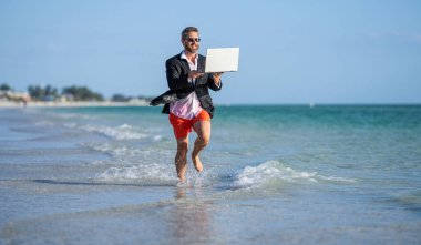 Başarılı bir iş adamı yazın tadını çıkarır. Yaz tatilindeki iş adamı. İnsanoğlunun iş başarısı. Denizde dizüstü bilgisayarla koşan takım elbiseli bir adam. Sıcak yaz işleri..