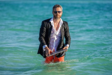 İş adamı takım elbiseleri sahilde rahatlar. Resmi olarak dizüstü bilgisayarı olan iş adamı tropik plaj yakınlarında giyiniyor. Sıcak iş yazı. İş patronu haftasonunu sahilde geçiriyor..