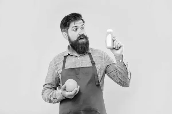 stock image Surprised man in apron holding orange and juice bottle yellow background, bartender.