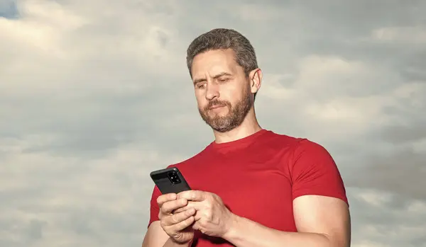 stock image man blogger typing phone message outdoor. man blogger type phone message wear red tshirt. photo of man blogger type phone message. man blogger type phone message on sky background.