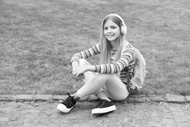 Happy teen girl with school backpack listening to music sitting on grass outdoors, education.