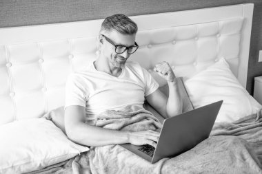 Gözlüklü mutlu bir adam laptopla yatakta çalışıyor..