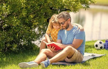 Baba ve çocuk kitap okuyor. Baba ve oğul açık havada okuyorlar. Baba ve oğul doğada edebi maceralar yaratır. Baba oğul dışarıda okuma zamanı. Baba ve oğul için kutsal bir gelenek. aile açık hava eğitimi.