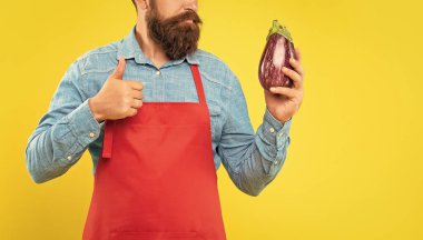 Serious man crop view in red apron giving thumb gesture to eggplant yellow background, greengrocer. clipart