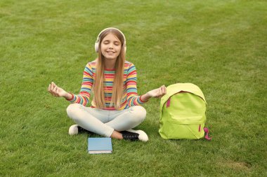 Meditating girl listening to music with closed eyes sitting on grass after school, education.