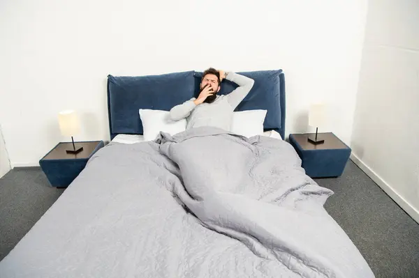 stock image Sleepy guy yawning and stretching in bed waking up after sleep in bedroom, morning.