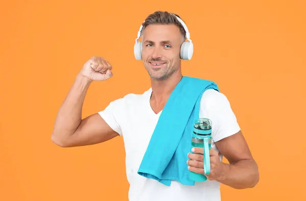 stock image successful fitness man holding sport bottle in studio. fitness man with towel and bottle after training. fitness man in headphones hold sport bottle. fitness man with sport bottle isolated on yellow.