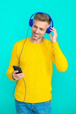 Telefonla müzik dinlemek. Müzik yaşam tarzı. Ses çalma listesi. Kulaklık takan olgun bir adam. Kulaklıklı adam telefonda çalma listesini seçiyor. Telefon uygulaması kullanan adam mavi ile izole edilmiş. Müzikle spor yapmak.