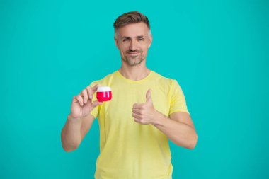 Mavi renkte yüz kremi gösteren bir adam var. Olgun adam cilt kremi kullanır. Günlük alışkanlık ve kişisel bakım. Cilt güzelliği. Erkek güzellik kozmetiği. Erkek kozmetik ürünü sunan adam. Başparmak yukarı..