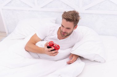 Time to wakeup and get up. Morning wakeup. Sleepy man looking at alarm clock in hand while lying in bed. Morning awakening. Sleepy man turning off alarm clock. Waking up early. Turns off Alarm Clock. clipart