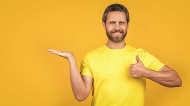 Arka planda reklam sunan mutlu adam, başparmağını kaldır. Stüdyoda reklam sunan adam. Reklam sunan adamın fotoğrafı. Reklam sunan adam sarı üzerine izole edilmiş..