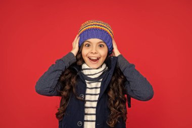 Şapkalı, kıvırcık saçlı, hayret verici çocuk. Kırmızı arka plandaki genç kız. Kalın giysiler giyen çocuk portresi. Olumlu duygularını ifade et. kış modası.