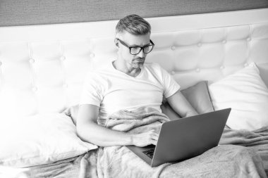 Yatakta dizüstü bilgisayarda çalışan gözlüklü yetişkin işveren..