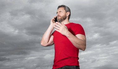photo of man has phone talk, communicate. man has phone talk on sky background. man has phone talk outdoor. man has phone talk wear red tshirt.