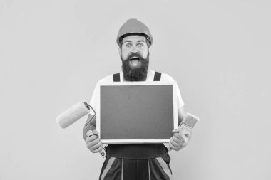 amazed man in helmet and work clothes hold paint roller brush and blackboard with copy space on yellow background.