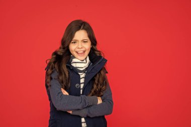 emotional kid with curly hair in puffer waistcoat. teen girl on red background. portrait of child wearing warm clothes. express positive emotion. winter fashion. copy space.