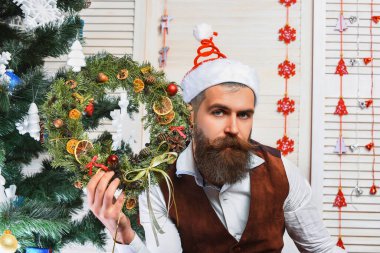 Bıyıklı adam yılbaşı çelengini Noel Baba 'nın Noel şapkasında evde otururken takıyor..