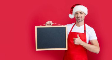 Noel şapkalı ve önlüklü gülümseyen olgun bir adam fotokopi alanı olan karatahtayı tutuyor. başparmak yukarı.