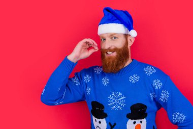 Kafkasyalı mutlu adam Noel süveteri ve Noel Baba şapkası ile kırmızı arka planda izole edilmiş fotokopi alanı ile. Adam Noel 'i kutluyor. Mutlu Noeller..