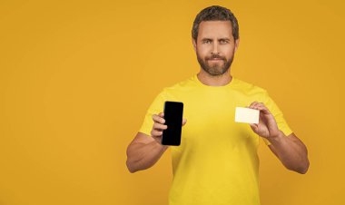 Stüdyoda akıllı telefon ve kart gösteren bir adam, afiş. Akıllı telefon ve kart gösteren bir adamın fotoğrafı. Akıllı telefon ve kart gösteren adam sarıda izole edilmiş. Adam arka planda akıllı telefon ve kart gösteriyor.