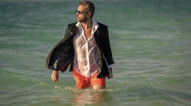 İş adamı takım elbiseleri sahilde rahatlar. Resmi olarak dizüstü bilgisayarı olan iş adamı tropik plaj yakınlarında giyiniyor. Sıcak iş yazı. İşletme patronu hafta sonunu sahilde geçiriyor. seyahat ve rahatlama.