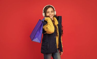 happy child in winter clothes and headphones hold shopping bag on red background, sale.