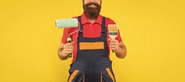 İş kıyafetleri içinde kırpılmış adam dekoratörü. Sarı arka planda boya silindiri ve fırça tutuyor..