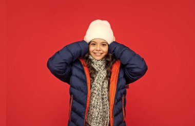 Kış modası. Kabarık ceketli ve şapkalı, kıvırcık saçlı sevimli çocuk. Kırmızı arka plandaki genç kız. Eşarplı sıcak kıyafetler giyen bir çocuğun portresi. pozitif duygularını ifade et.