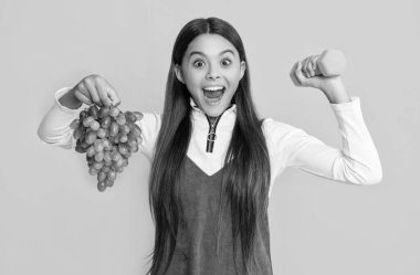 amazed smiling child hold fresh grapes and sport barbells on yellow background.