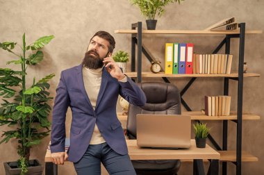 Takım elbiseli ciddi bir çalışan cep telefonuyla konuşuyor. Ofis masasında oturuyor, iletişim kuruyor..