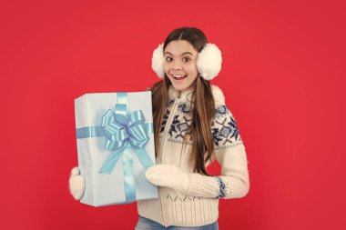 Noel tatili. Kış kulaklıklarıyla yılbaşı hediyesiyle hayrete düşmüş genç bir kız. Genç kız Noel hediyesi olarak kırmızı arka planda izole edilmiş..