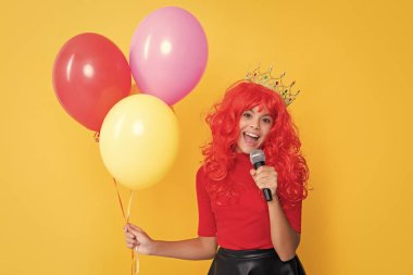 Sarı arka planda parti balonu ve mikrofonla taç takmış çocuk gülümsemesi..