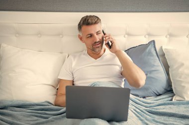Olgun bir adam telefonda konuşur ve laptopla yatakta çalışır..