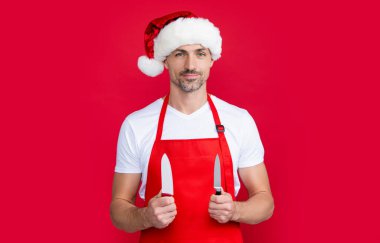 Kırmızı Noel Baba şapkalı ve önlüklü olgun bir adam yemek bıçağını tutar..