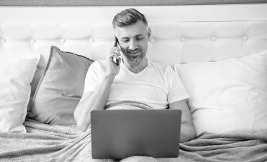 Neşeli, olgun bir adam telefonda konuşuyor ve laptopla yatakta çalışıyor..