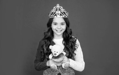 Kraliçe tacındaki mutlu çocuk. Tacın içindeki prenses. Çocuk ayı oyuncağını tutuyor. Genç kız kırmızı arka planda diyadem giyiyor. Mutlu çocukluk.